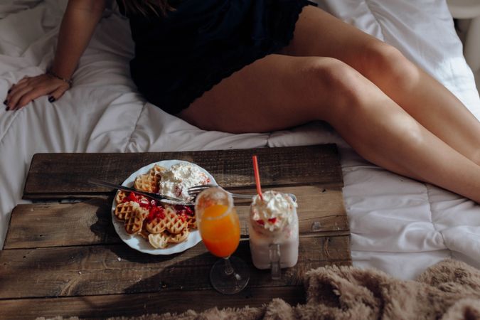 Young woman eating appetizing waffles and tasty drinks on tray on bed