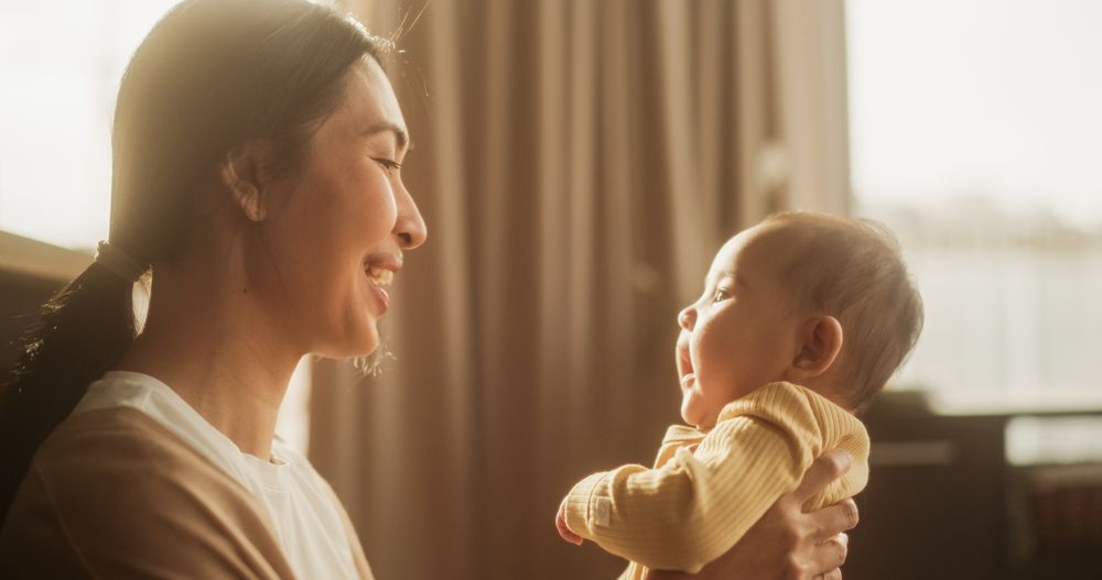 Close Up Portrait of an Affectionate Asian Mother Smiling and Playing with her Baby in a Cosy Naturally Lit Bedroom.