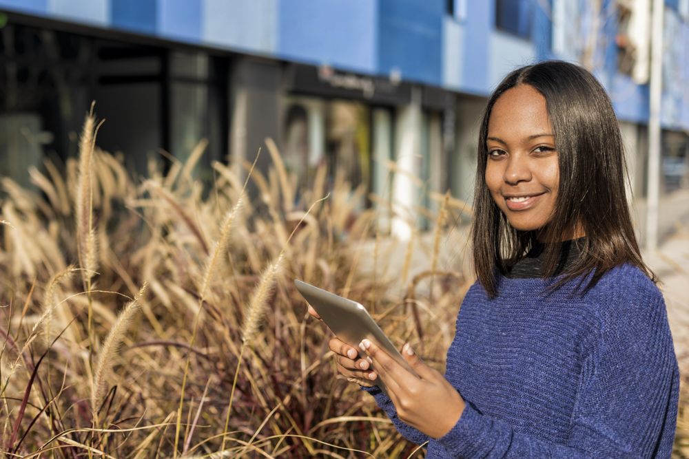 Confident female in sweater standing outside blue building with tablet