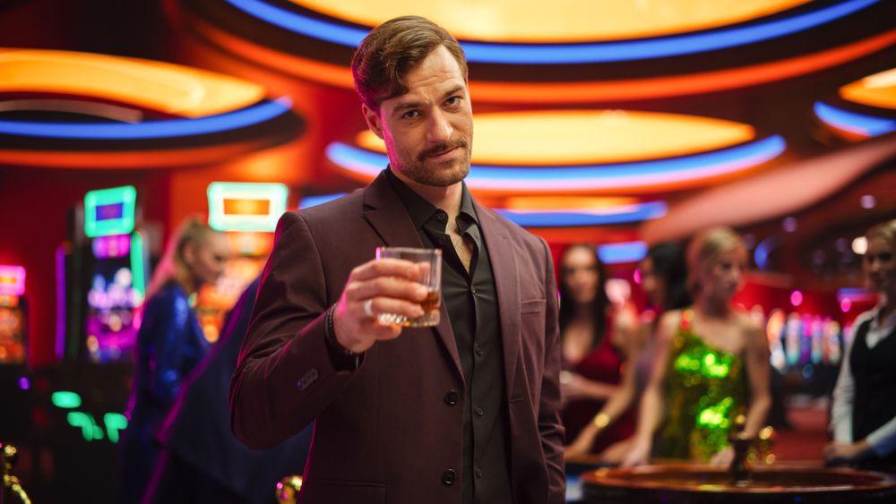 Portrait of a Sophisticated Young Man in a Suit, Smiling, Looking at Camera and Raising a Whisky Glass in a Luxurious Casino