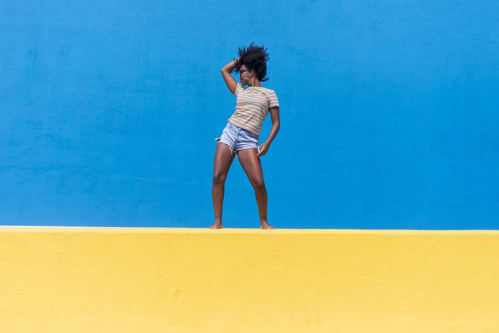 Black woman posing atop bright yellow and blue wall