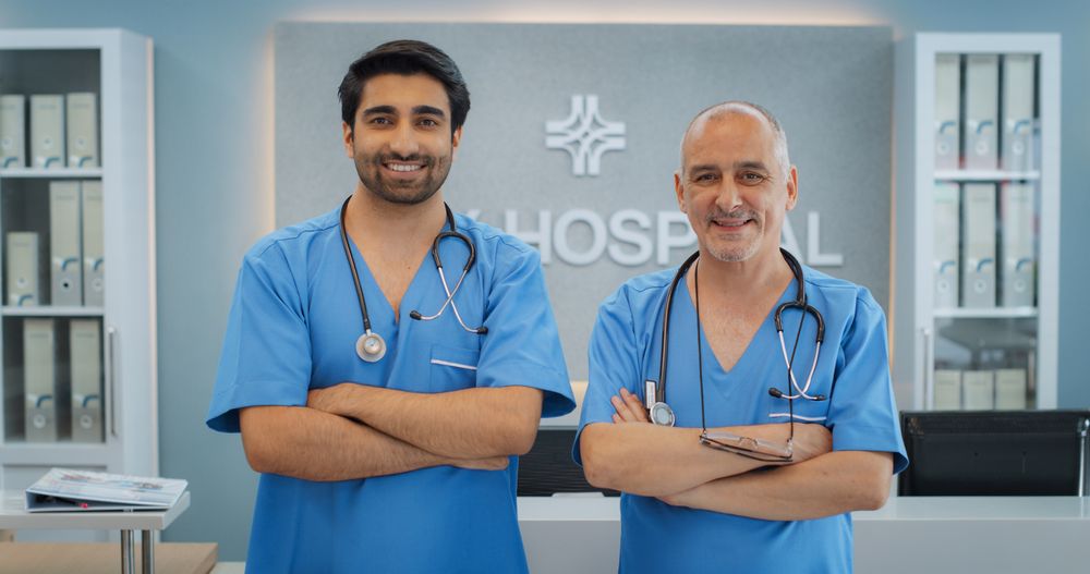 Two Friendly Indian Male Medical Doctors Crossing Their Hands, Looking at Camera and Smiling.