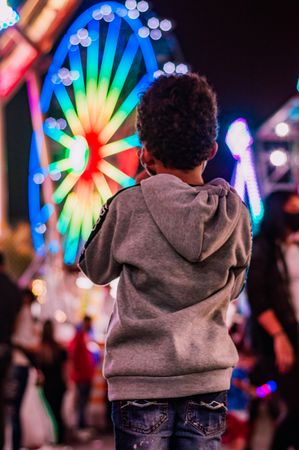 Young boy is grey jacket standing in front of colorful Ferris wheel