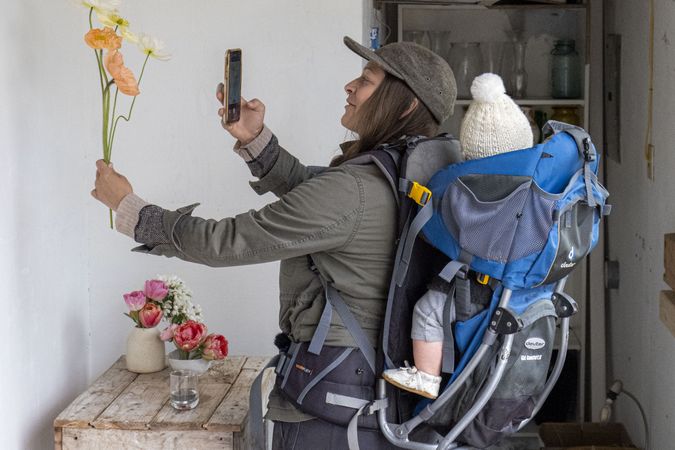 Woman with baby on back taking picture of flower with phone
