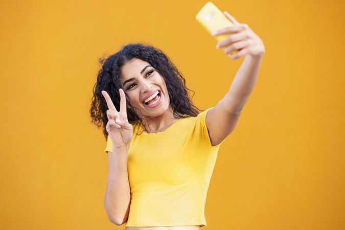 Female in yellow t-shirt taking selfie making peace sign in front of mustard background