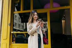 Young cheerful female standing near house with smartphone