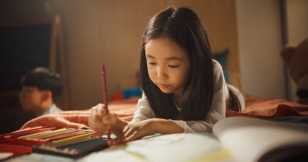 Portrait of a Little South Korean Girl Drawing with Colored Pencils in a Notebook While Lying in Bed in Children's Bedroom at Home.