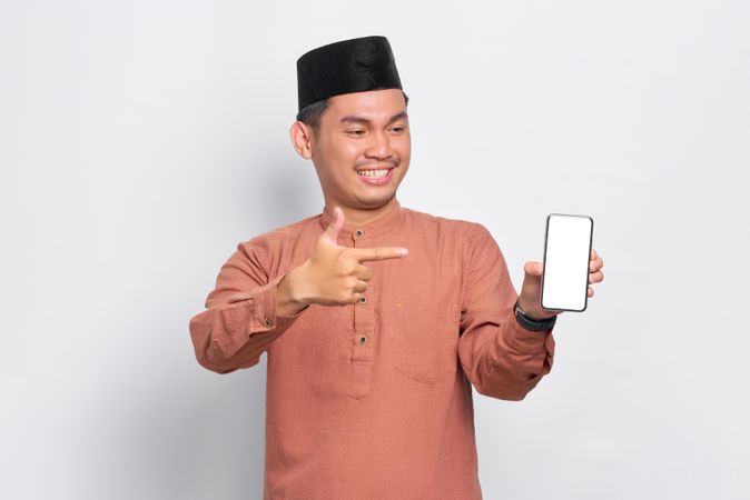 Muslim man in kufi hat smiling and pointing at blank screen on mobile phone