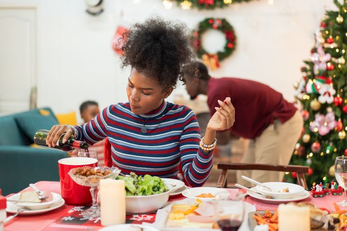 Black woman is pouring red wine into a glass at Christmas dinner table