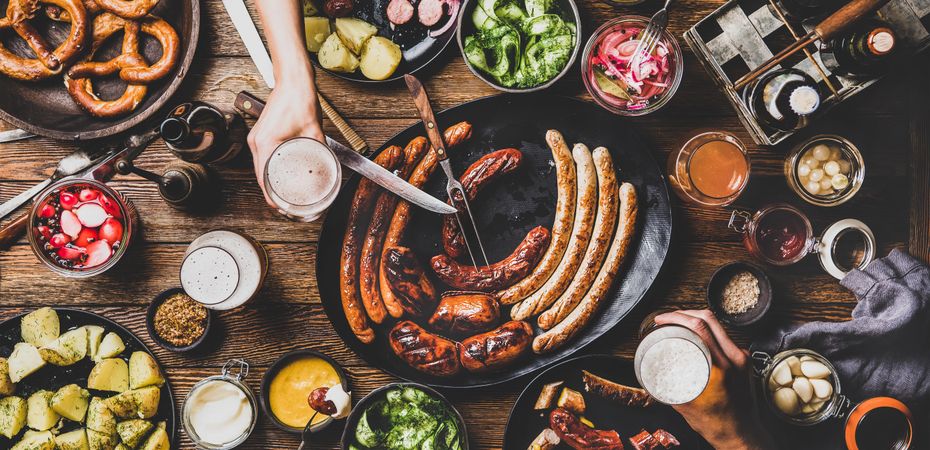 German sausages and pretzels displayed on wooden table with hands holding beer