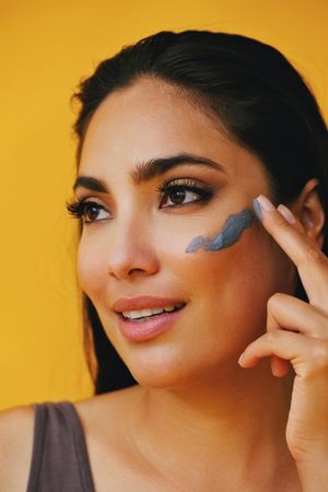 Latina woman looking away while applying clay mask with hand, vertical