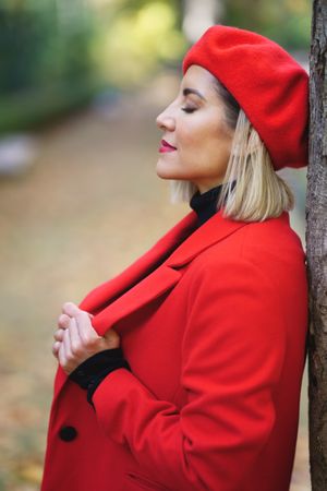 Side view of peaceful female in red coat and beret standing with closed eyes near tree trunk in autumn park on blurred background