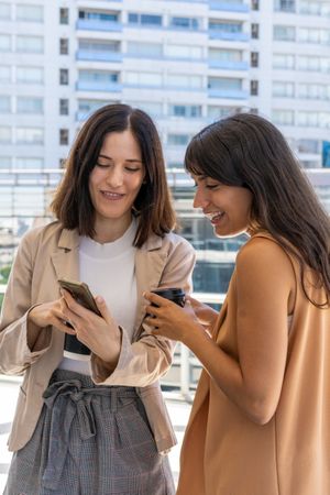 Two women are outside. One shows something on her phone while the other holds a cup