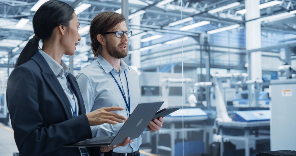 Portrait of an Experienced Male and Female Engineers Standing in Agency Room, Using Computer Devices and Discussing Production at a Modern Automated Electronics Manufacture with Robot Arms