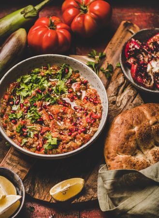 Eggplant dip with pomegranate and parsley garnish on wooden surface with vegetables and bread