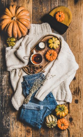Flat-lay of beige knitted sweater, blue jeans, hat, decorative pumpkins, candle and tea