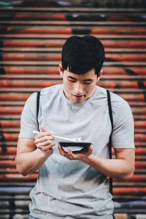 Asian man eating takeaway food with chopsticks on city street