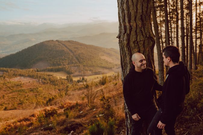Smiling men hugging near tree in forest on hill