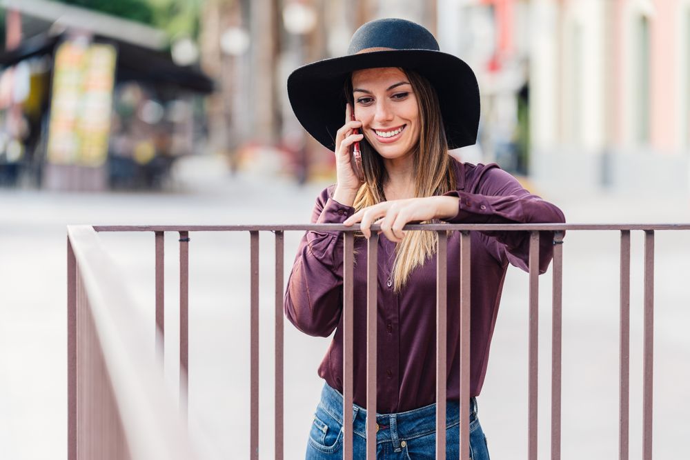 Charming woman in trendy outfit talking on smartphone and smiling at city street