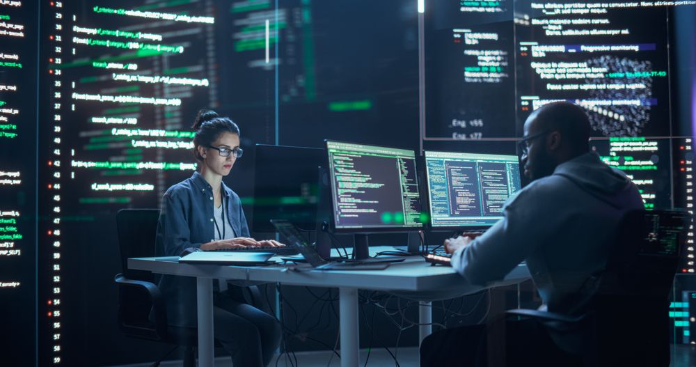 Portrait of Two Programmers Working in a Monitoring Control Room, Surrounded by Big Screens Displaying Lines of Programming Language Code.