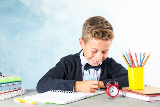 Boy in school uniform writes sitting at the table against color background