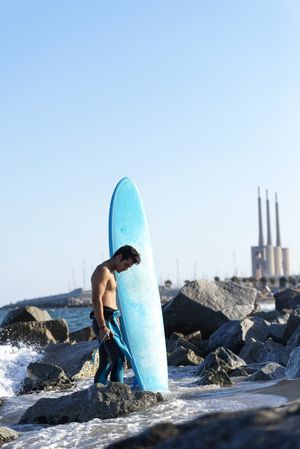 Male surfer with blue board standing in the water around the rocky shore