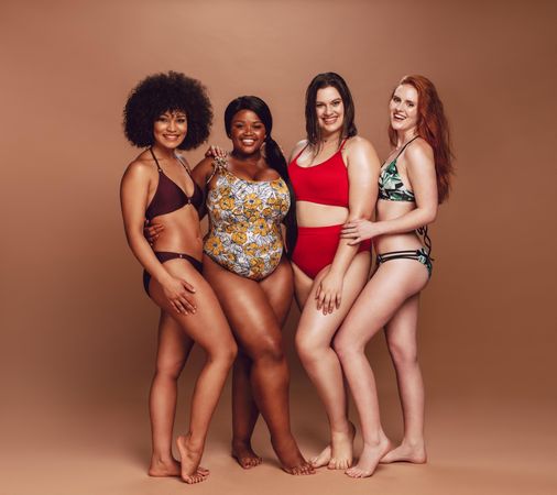 Full length of multiracial women in swimsuits looking at camera and smiling