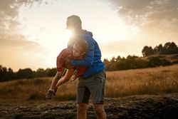 A father playfully lifting his daughter in the air like an airplane at sunset.