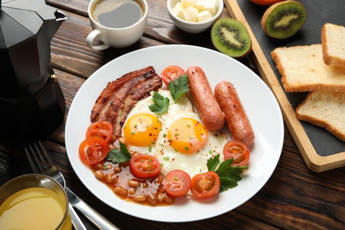 Wooden table with breakfast plate of coffee eggs, tomatoes, sausage and bacon, top view