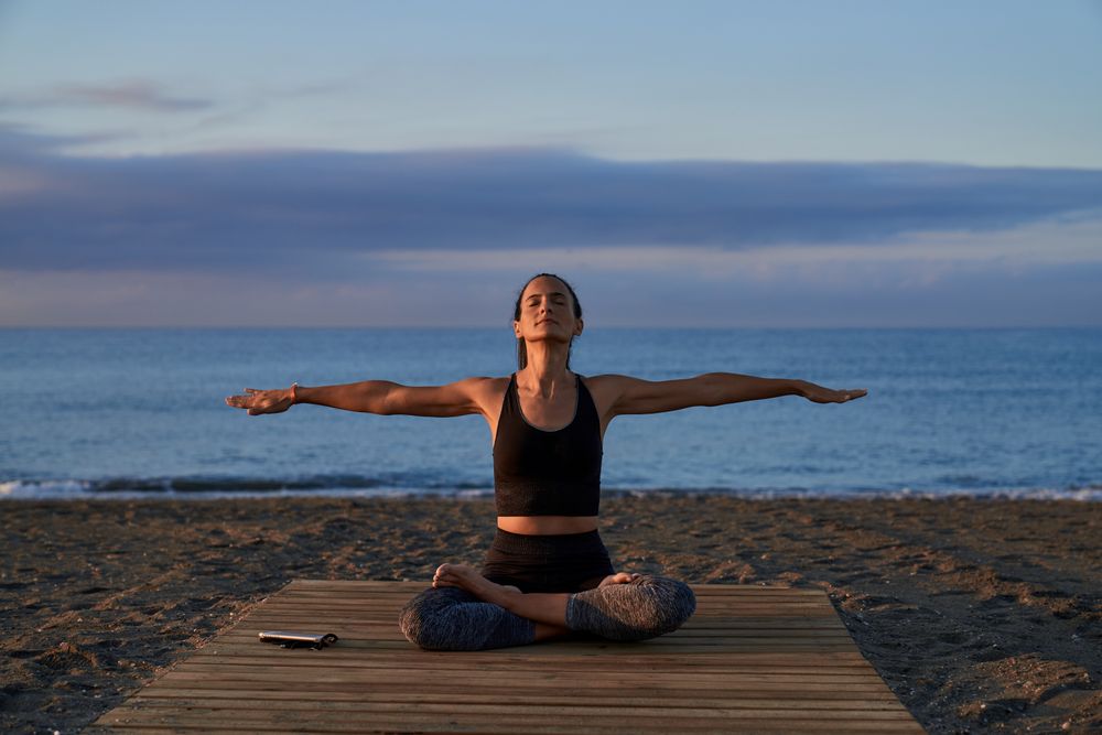 Hispanic woman in lotus pose with spread arms on shore