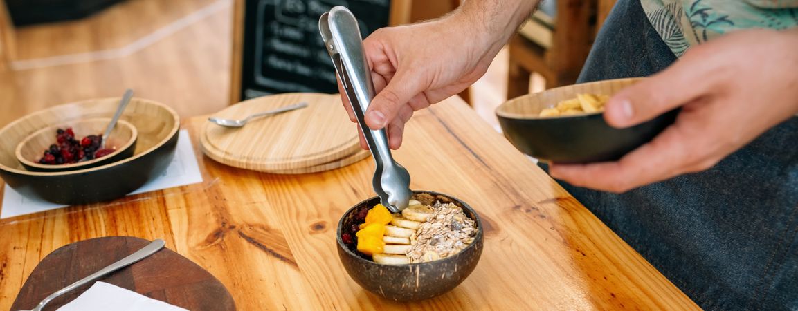 Chef preparing vegan breakfast with mango, banana, raspberries, cereals and muesli in coconut bowl