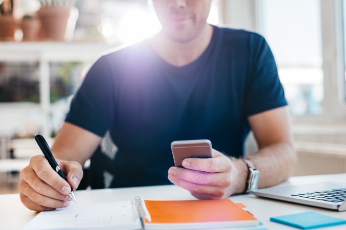 Man in business casual clothes  using mobile phone while writing in notebook