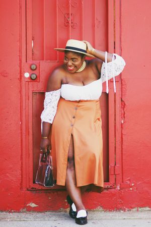 Woman in light shirt and orange skirt wearing hat standing beside red door