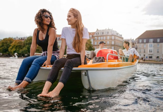 Shot of two women sitting in front pedal boat with feet in water and man in background