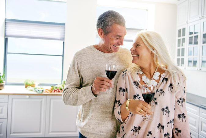 Grey haired older couple smiling at each other in the kitchen while holding red wine