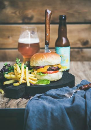 Classic hamburger skewered with knife with fries and beer bottle at wooden restaurant table
