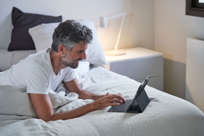 Mature man typing on tablet keyboard while lying on bed at home