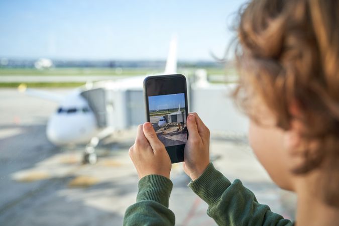 Crop kid photographing airplane on smartphone through window in