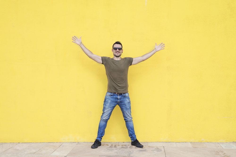 Happy male standing outside in front of yellow wall with open arms
