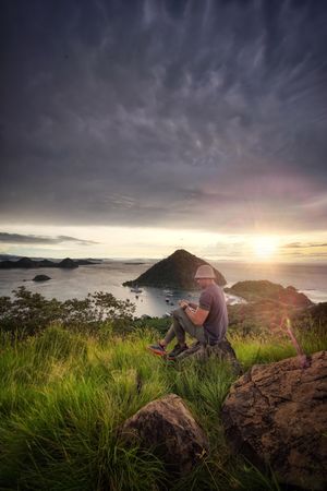 Back view of young man sitting near seashore at sunset in Sylvia Hills - Labuan Bajo, East Nusa Tenggara, Indonesia