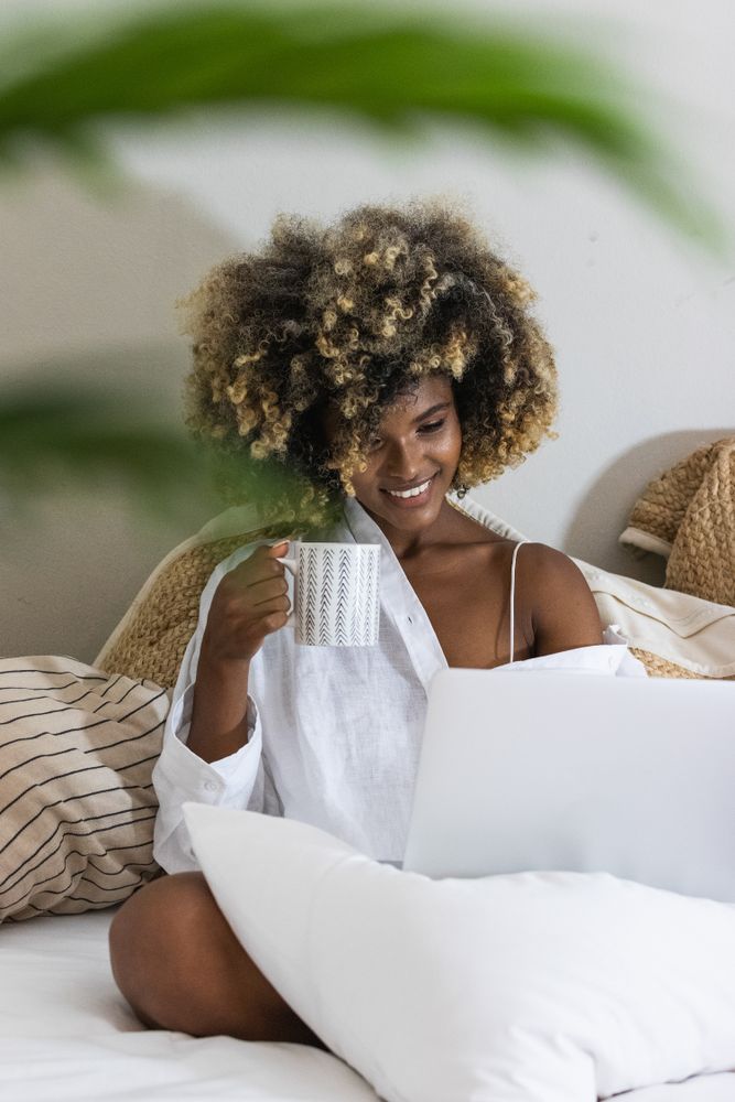 Black woman drinking coffee while sitting on bed with laptop