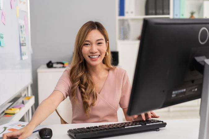 Happy Asian woman sitting in office behind computer and monitor