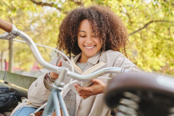 Positive Black Latina woman reading book on bench in park