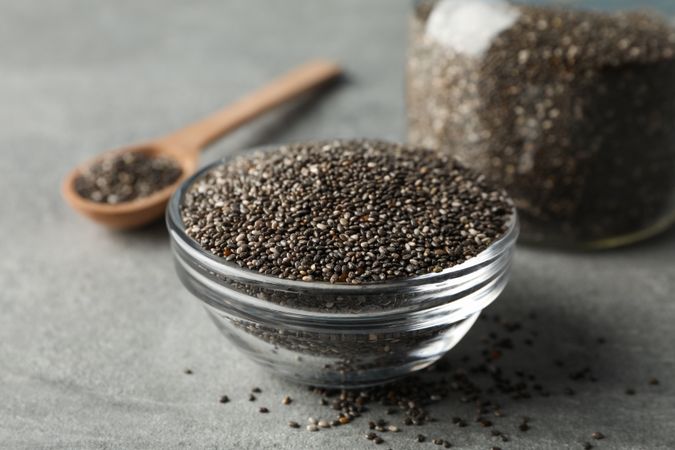 Glass bowl, jar and wood spoon with chia on grey background, closeup