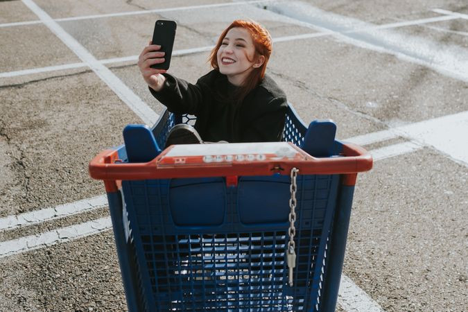Smiling woman with smartphone in shopping trolley in parking lot