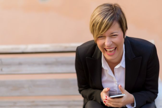 Laughing woman in blazer sitting on bench holding phone in front of peach wall