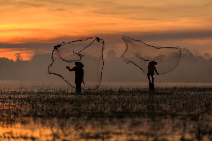 Silhouette of two fishermen with conical hat throwing their fishing net into water at sunset