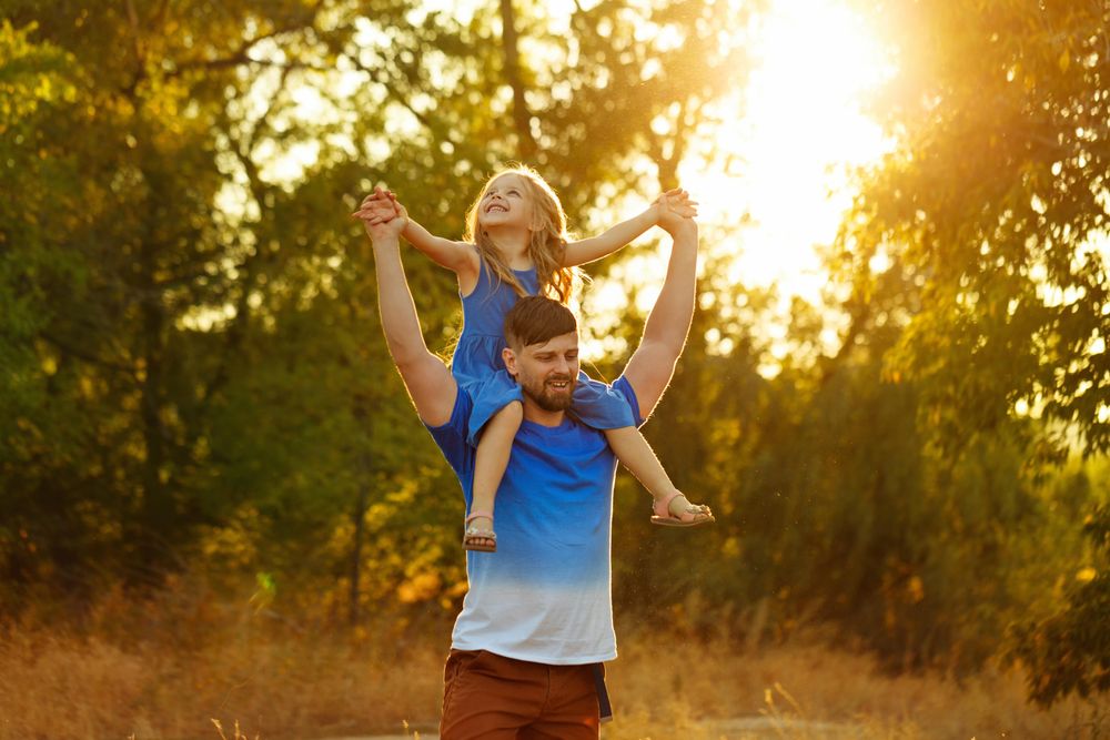 Man with daughter on shoulders dressed in blue walking along forest trail