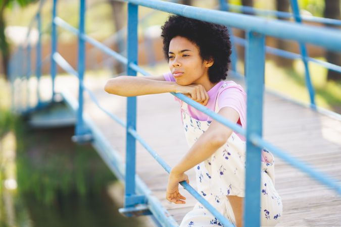 Woman in floral overalls sitting on bridge over water deep in thought