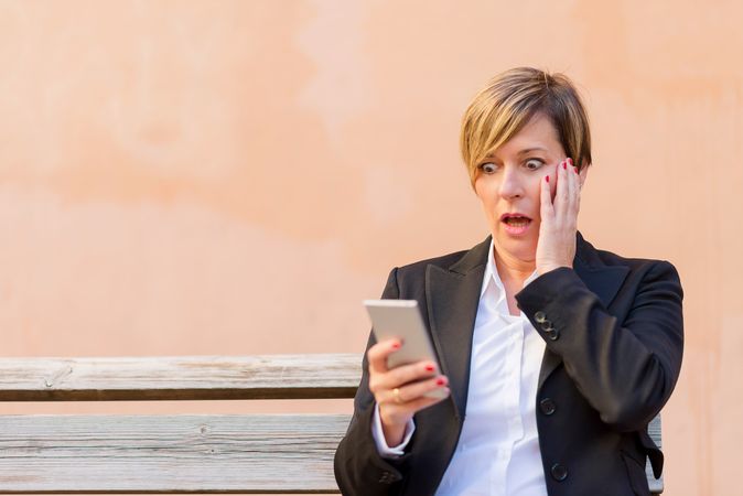 Shocked female in blazer sitting on bench checking phone in front of peach wall